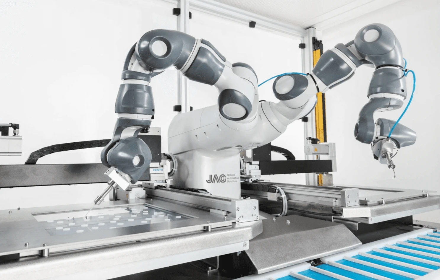 Dual-arm industrial robot performs precision pick-and-place on a tray of small parts inside a clean, enclosed workstation. Articulated arms with grippers and pneumatics operate on linear rails, automating assembly in a modern factory line.