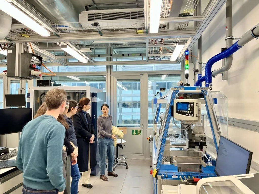 A small group listens to a presenter during a lab tour, standing beside a blue automated machine and computer workstations. Bright industrial lighting, cables, and ventilation indicate a modern research facility focused on robotics or automation.