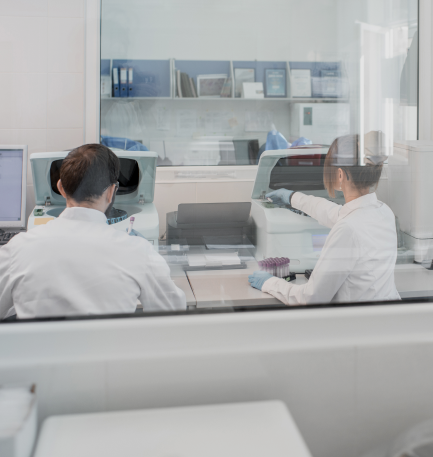 Two lab technicians in white coats operate laboratory equipment, analyzing samples in a clean, clinical lab environment seen through a glass partition.