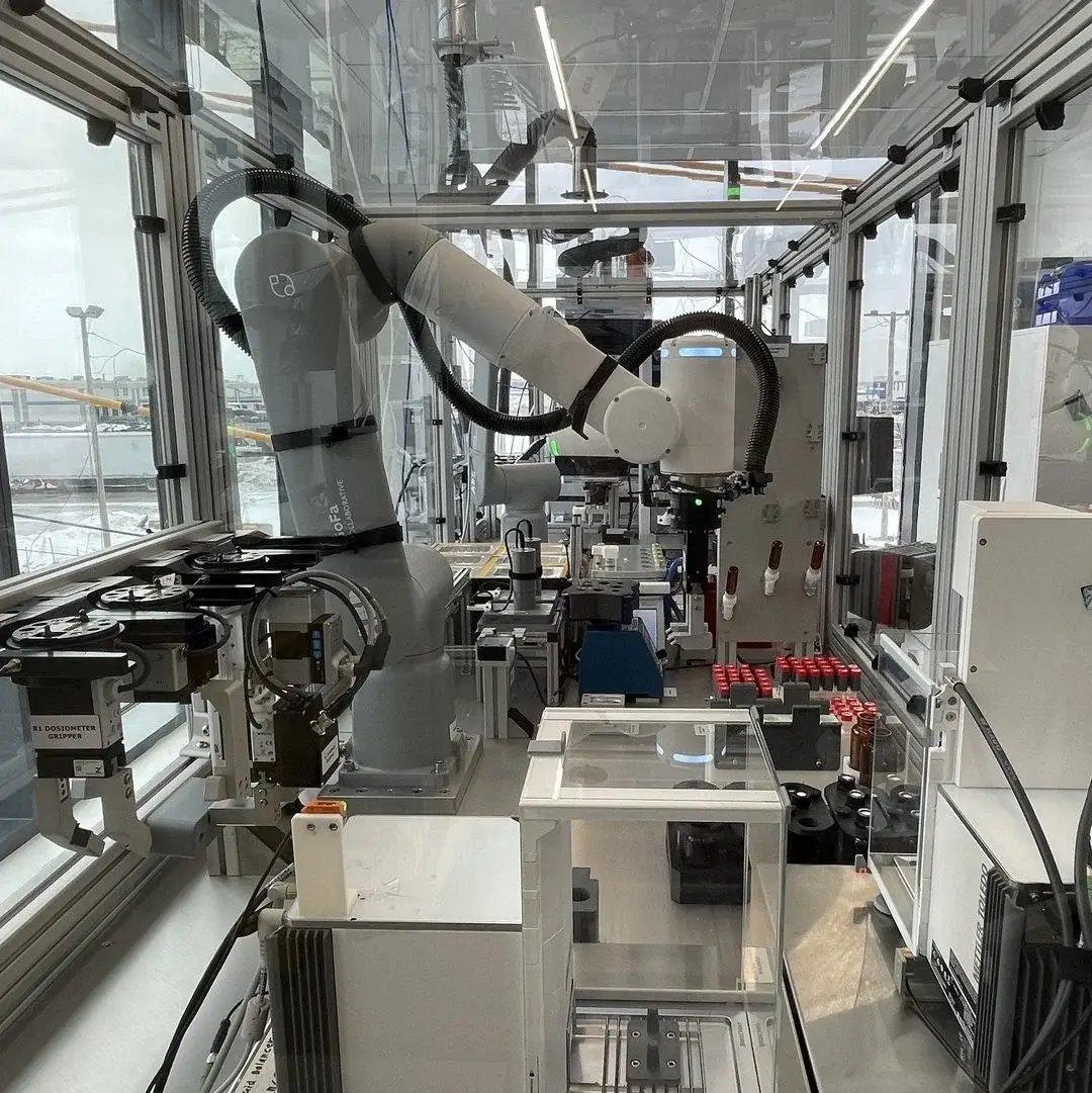 Enclosed laboratory automation cell with a robotic arm manipulating equipment and racks of red-capped vials. Sensors, grippers, and conveyors line the workstation inside a glass enclosure in a modern industrial research facility. This is showcasing Atinary's Self Driving Labs in Boston.