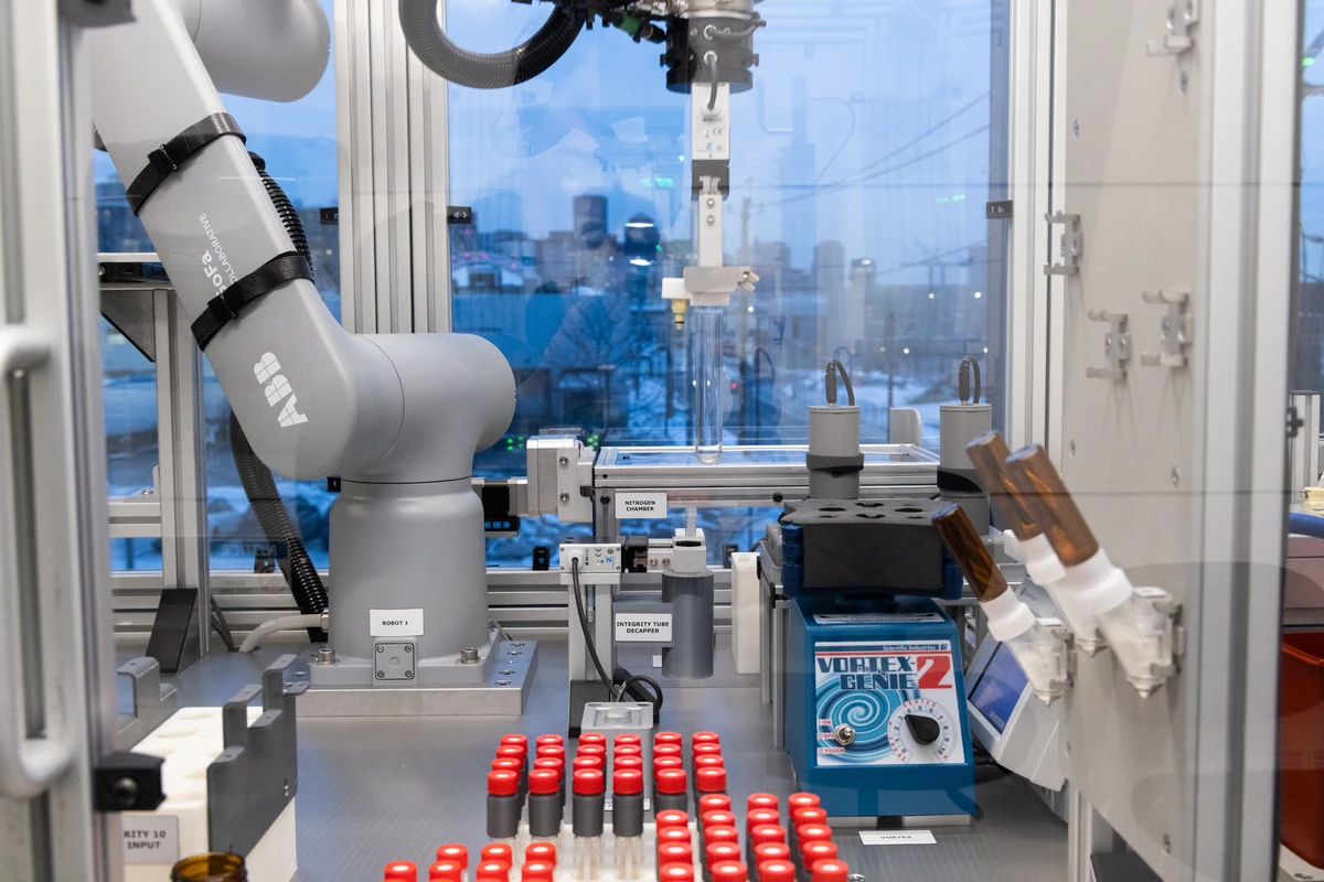 Enclosed laboratory automation station with ABB robotic arm picking a test tube utilizing SDLabs and Self Driving Labs capabilities. Benchtop includes racks of red-capped vials, an integrity tube decapper, and a Vortex Genie mixer, indicating automated sample handling.