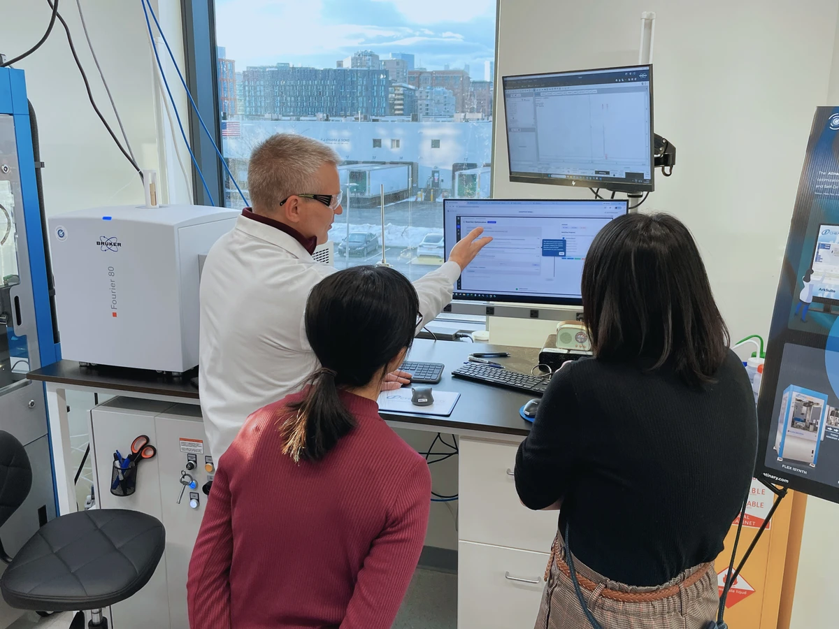 Three scientists in a laboratory; an instructor in a lab coat points to the SDLabs platform on a monitor while two scientists watch. Scientific instruments and keyboards surround the workstation, with city buildings visible through large windows.